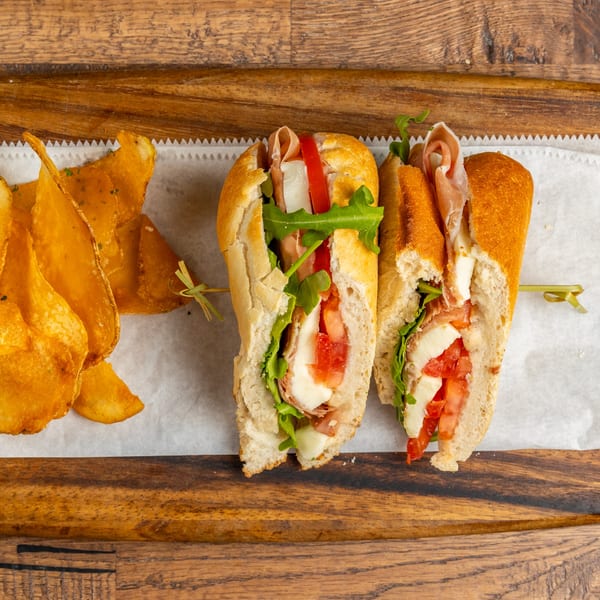 Prosciutto and mozzarella sandwich with tomatoes and arugula, served with potato chips on a wooden board.