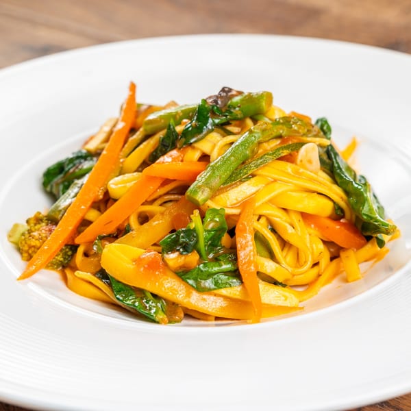 Close-up of a vibrant pasta dish with tagliatelle, carrots, green beans, and spinach on a white plate.