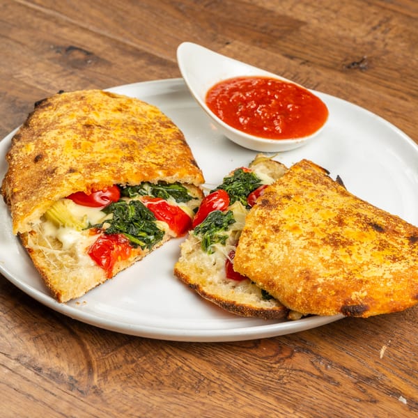 Two halves of a golden-brown calzone filled with spinach, tomatoes, and melted cheese, with a side of marinara.