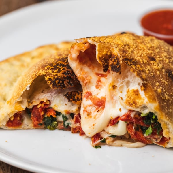 A golden-brown calzone from Tutto Pizza & Pasta, sliced open to reveal melted cheese, spinach, and sun-dried tomatoes.