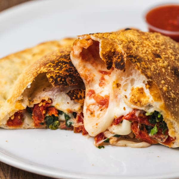 A golden-brown calzone from Tutto Pizza & Pasta, sliced open to reveal melted cheese, sun-dried tomatoes, and spinach.