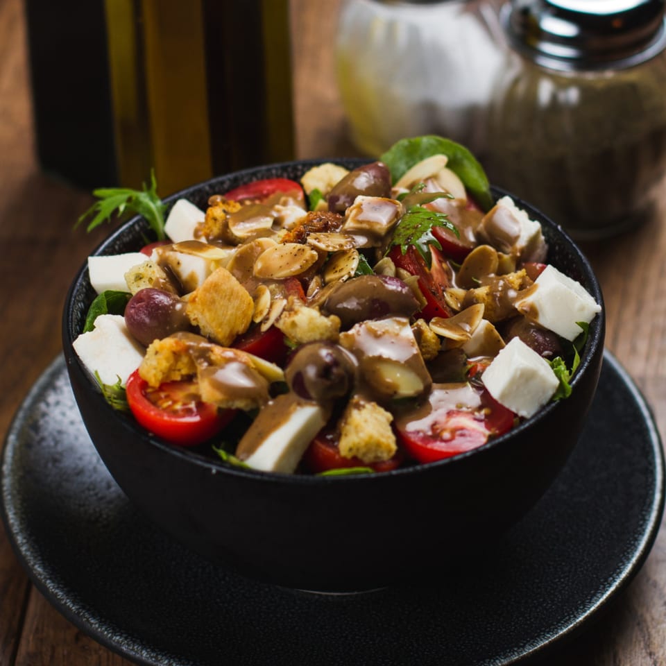 A vibrant salad with cherry tomatoes, feta, olives, croutons, and dressing in a black bowl.